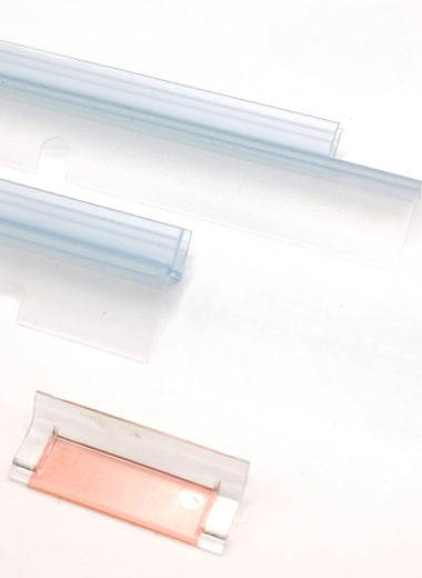 Nahaufnahme Ablaufprofil-Set der Marke HÜPPE bestehend aus zwei, transparenten, schmalen, länglichen, U-förmigen Kunststoffprofilen, mit einem Ablaufschutz an der Profilunterseite, in zwei Längen, sowie eine Kunststoffschiene, vor weißem Hintergrund.