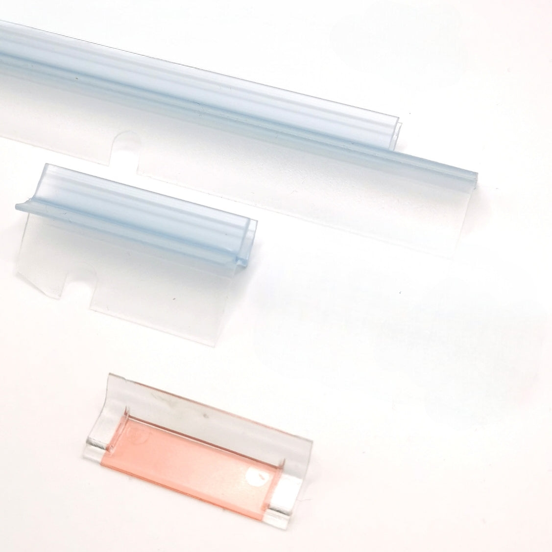 Nahaufnahme Ablaufprofil-Set der Marke HÜPPE bestehend aus zwei, transparenten, schmalen, länglichen, U-förmigen Kunststoffprofilen, mit einem Ablaufschutz an der Profilunterseite, in zwei Längen, sowie eine Kunststoffschiene, vor weißem Hintergrund.