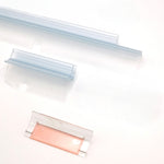 Nahaufnahme Ablaufprofil-Set der Marke HÜPPE bestehend aus zwei, transparenten, schmalen, länglichen, U-förmigen Kunststoffprofilen, mit einem Ablaufschutz an der Profilunterseite, in zwei Längen, sowie eine Kunststoffschiene, vor weißem Hintergrund.