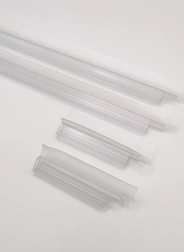 Nahaufnahme Ablaufprofil-Set der Marke HÜPPE, bestehend aus vier transparenten, schmalen, länglichen, U-förmigen Kunststoffprofilen, in zwei Längen, mit einem Ablaufschutz an der Profilunterseite bzw. Profilaußenkante, vor weißem Hintergrund.