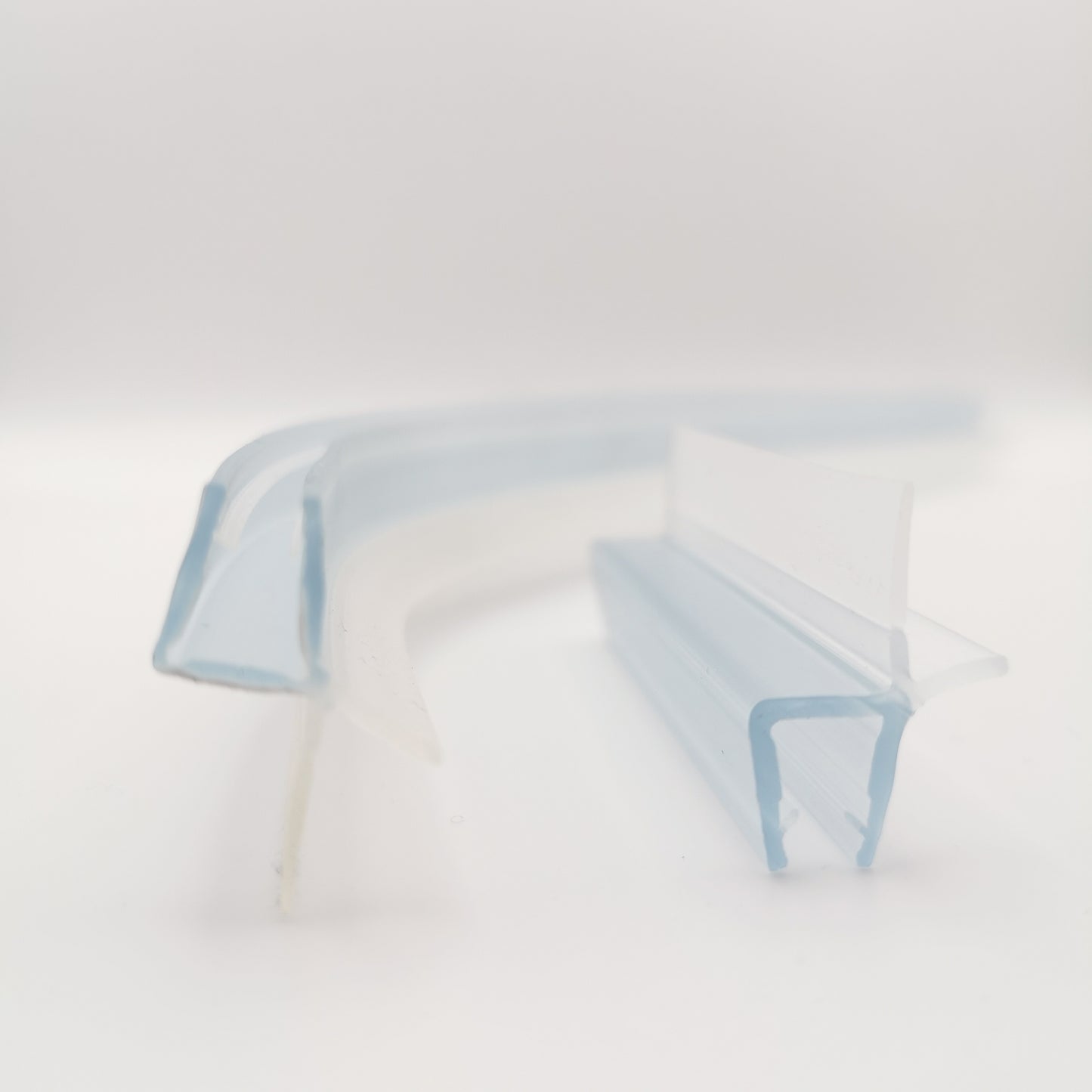 Detailaufnahme von zwei Ablaufprofilen der Marke HÜPPE, transparente, schmale, längliche, U-förmige Kunststoffprofile, mit einem Ablaufschutz an der Profilaußenkante, in zwei Längen sowie a-symmetrisch gebogen, vor weißem Hintergrund, Frontansicht.