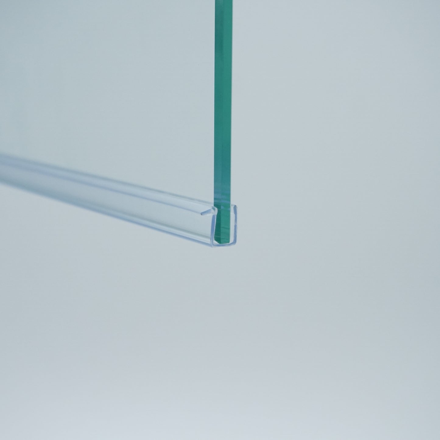 Nahaufnahme einer Ablaufleiste der Marke HÜPPE, transparentes, schmales, längliches, U-förmiges Kunststoffprofil, mit einem Vorsprung an der rechten Profiloberkante, montiert an der Unterseite einer Duschkabine, vor weißem Hintergrund, Seitenansicht links.