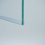 Nahaufnahme einer Ablaufleiste der Marke HÜPPE, transparentes, schmales, längliches, U-förmiges Kunststoffprofil, mit einem Vorsprung an der rechten Profiloberkante, montiert an der Unterseite einer Duschkabine, vor weißem Hintergrund, Seitenansicht links.