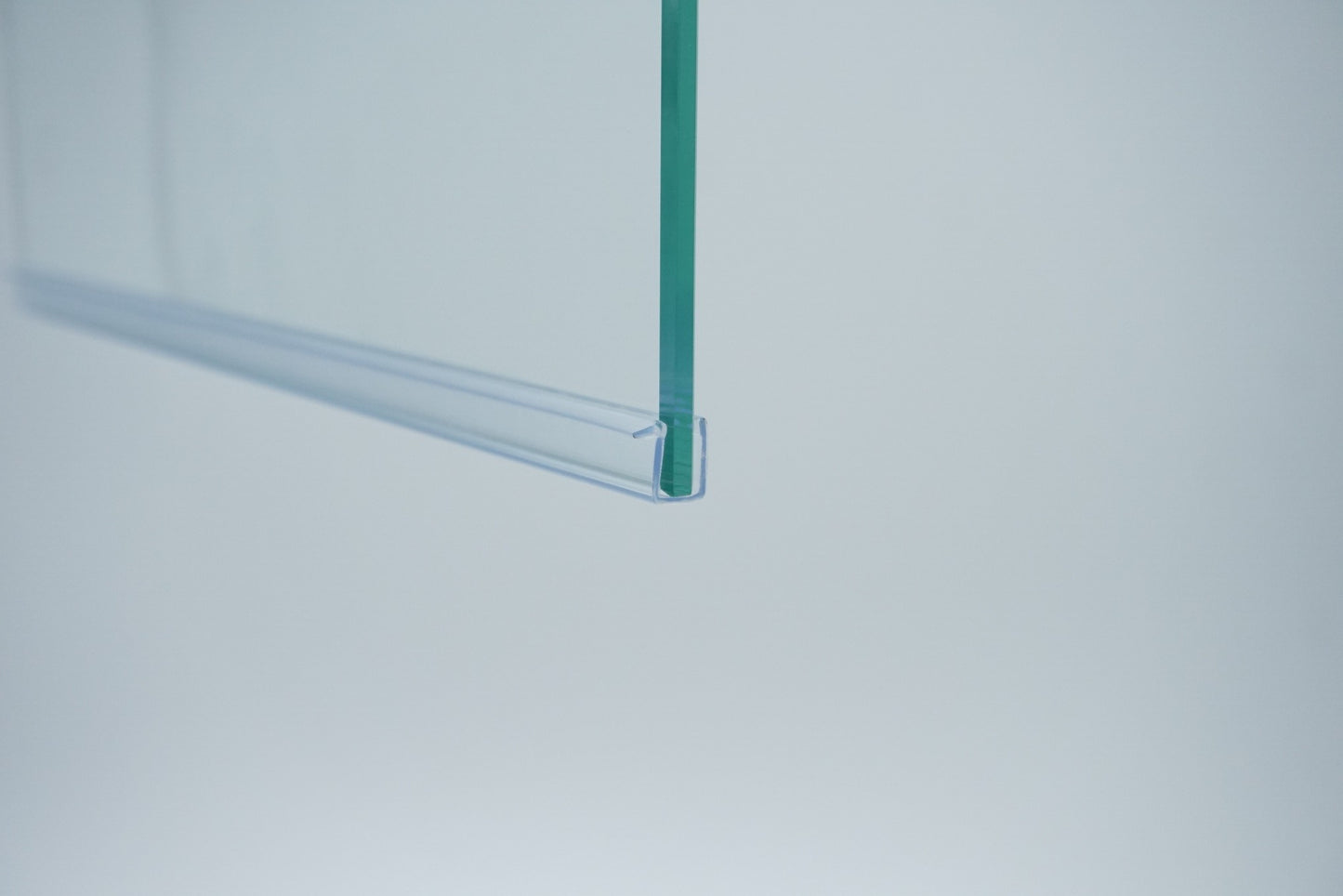 Nahaufnahme einer Ablaufleiste der Marke HÜPPE, transparentes, schmales, längliches, U-förmiges Kunststoffprofil, mit einem Vorsprung an der rechten Profiloberkante, montiert an der Unterseite einer Duschkabine, vor weißem Hintergrund, Seitenansicht links.