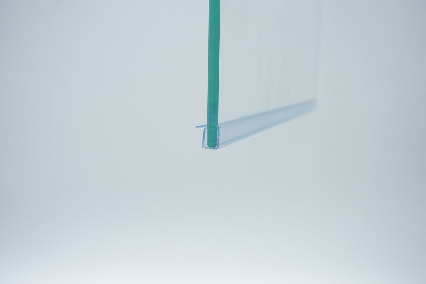 Nahaufnahme einer Ablaufleiste der Marke HÜPPE, transparentes, schmales, längliches, U-förmiges Kunststoffprofil, mit einem Vorsprung an der rechten Profiloberkante, montiert an der Unterseite einer Duschkabine, vor weißem Hintergrund Seitenansicht rechts.