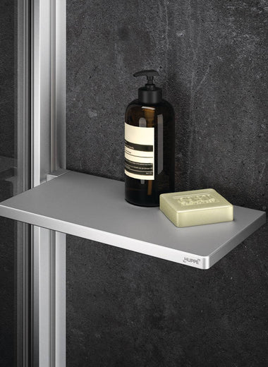 Select+ Tablet, Silberne Ablage der Marke HÜPPE befestigt an einer Wandleiste einer Glasdusche, darauf steht ein Seifenspender und eine Handseife, vor einer dunklen strukturierten Wand.