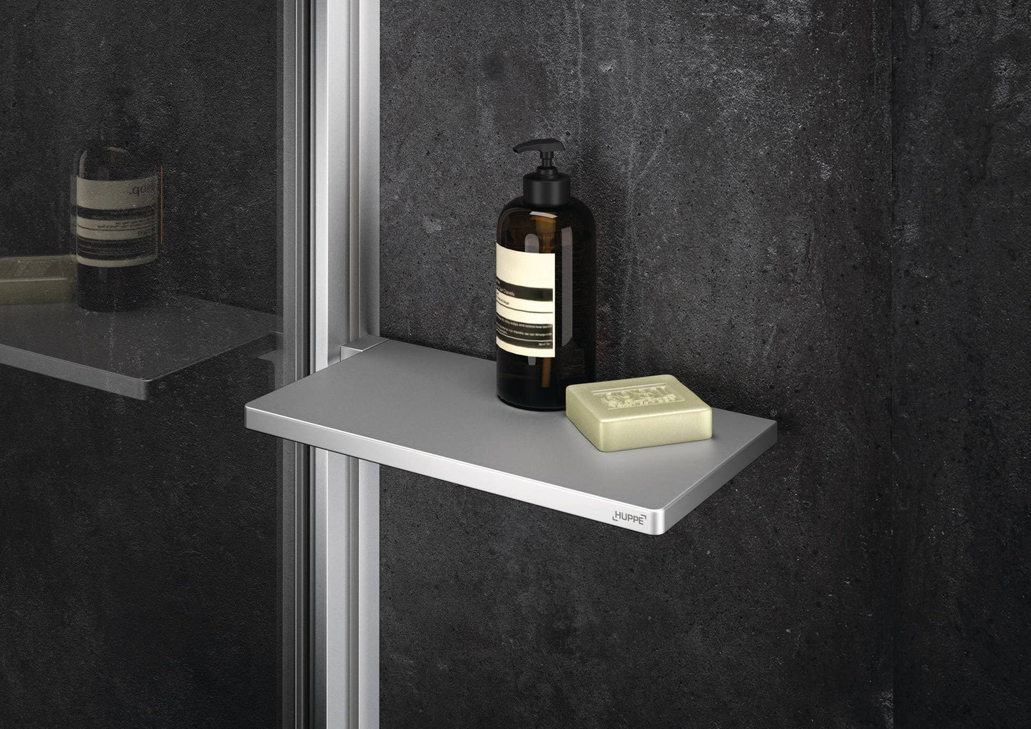 Select+ Tablet, Silberne Ablage der Marke HÜPPE befestigt an einer Wandleiste einer Glasdusche, darauf steht ein Seifenspender und eine Handseife, vor einer dunklen strukturierten Wand.