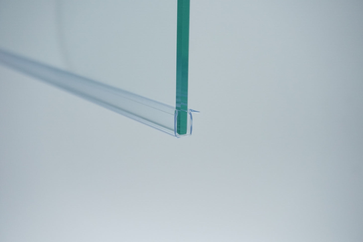 Nahaufnahme einer Ablaufleiste der Marke HÜPPE, transparentes, schmales, längliches, U-förmiges Kunststoffprofil, mit einem Vorsprung an der rechten Profiloberkante, montiert an der Unterseite einer Duschkabine, vor weißem Hintergrund, Seitenansicht links.