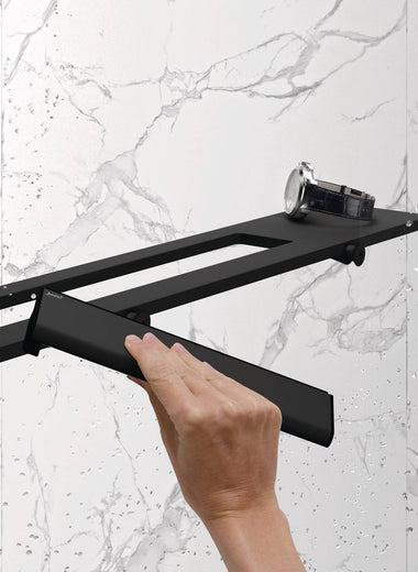 Hand wischt Wassertropfen von einer Glasdusche mit dem Select+ Wiper in schwarz matt, der Marke HÜPPE, während eine Uhr auf einer modernen Ablage vor einer hellen, strukturierten Wand liegt.