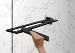 Hand wischt Wassertropfen von einer Glasdusche mit dem Select+ Wiper in schwarz matt, der Marke HÜPPE, während eine Uhr auf einer modernen Ablage vor einer hellen, strukturierten Wand liegt.