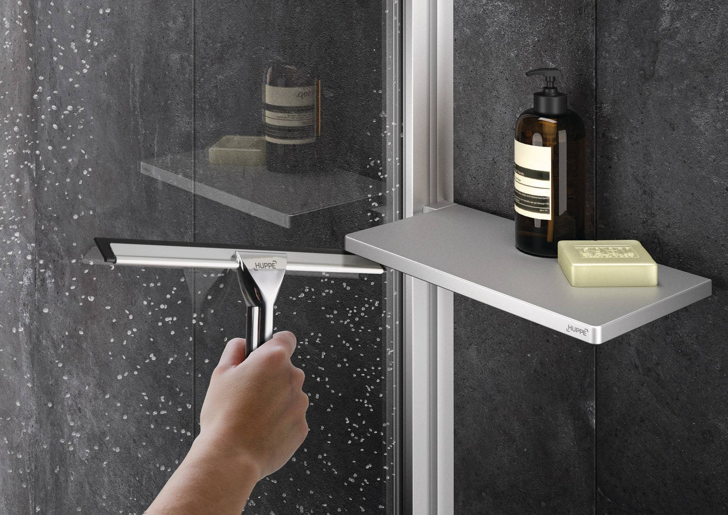 Select+ Tablet, Silberne Ablage der Marke HÜPPE befestigt an einer Wandleiste einer Glasdusche, darauf steht ein Seifenspender und eine Handseife, vor einer dunklen strukturierten Wand. Hand wischt Wassertropfen von einer Glasduschwand mit einem Silber glänzenden Handwischer trocken.