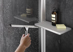 Select+ Tablet, Silberne Ablage der Marke HÜPPE befestigt an einer Wandleiste einer Glasdusche, darauf steht ein Seifenspender und eine Handseife, vor einer dunklen strukturierten Wand. Hand wischt Wassertropfen von einer Glasduschwand mit einem Silber glänzenden Handwischer trocken.