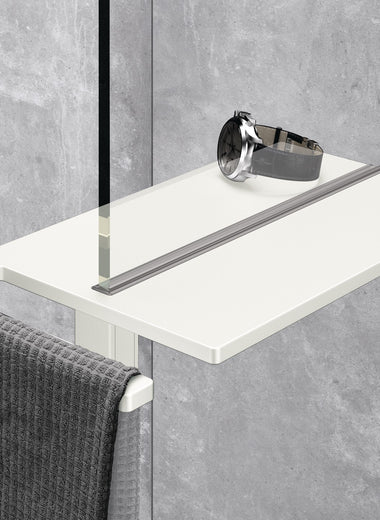 Select+ Showerboard, Klemmbares Ablagebrett der Marke HÜPPE in weiß matt, befestigt an einer Glasdusche, auf der eine Uhr und ein graues Handtuch abgelegt sind, vor einem hellgrauen, strukturierten Hintergrund.