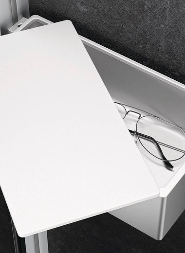 Silberne Select+ Box der Marke HÜPPE befestigt an einer Wandleiste einer Glasdusche, mit weißem, offenstehendem Deckel, darin liegt eine Brille, vor einer dunklen, strukturierten Wand.