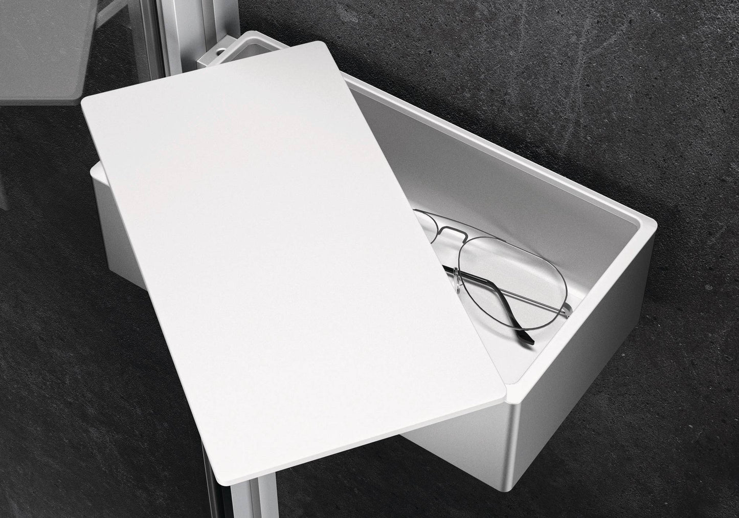 Silberne Select+ Box der Marke HÜPPE befestigt an einer Wandleiste einer Glasdusche, mit weißem, offenstehendem Deckel, darin liegt eine Brille, vor einer dunklen, strukturierten Wand.