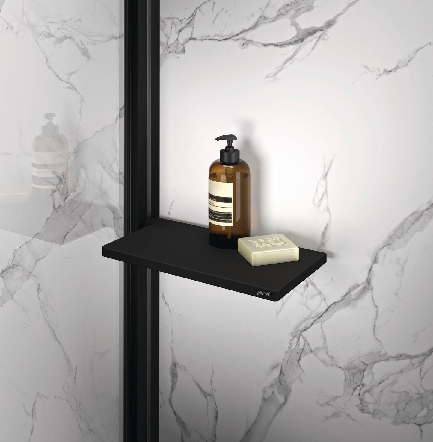 Select+ Tablet, Schwarze, matte Ablage der Marke HÜPPE befestigt an einer Wandleiste einer Glasdusche, darauf steht ein Seifenspender und eine Handseife, vor einer hellen strukturierten Wand.