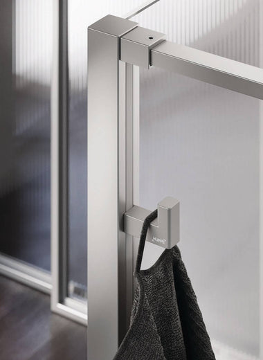 Select+ Hook, silberner Haken der Marke HÜPPE, der an einem senkrechten Profil einer Glasdusche montiert ist, daran hängt ein graues Handtuch, in einem modernen Badezimmer mit dunkelbraunem Fußboden.