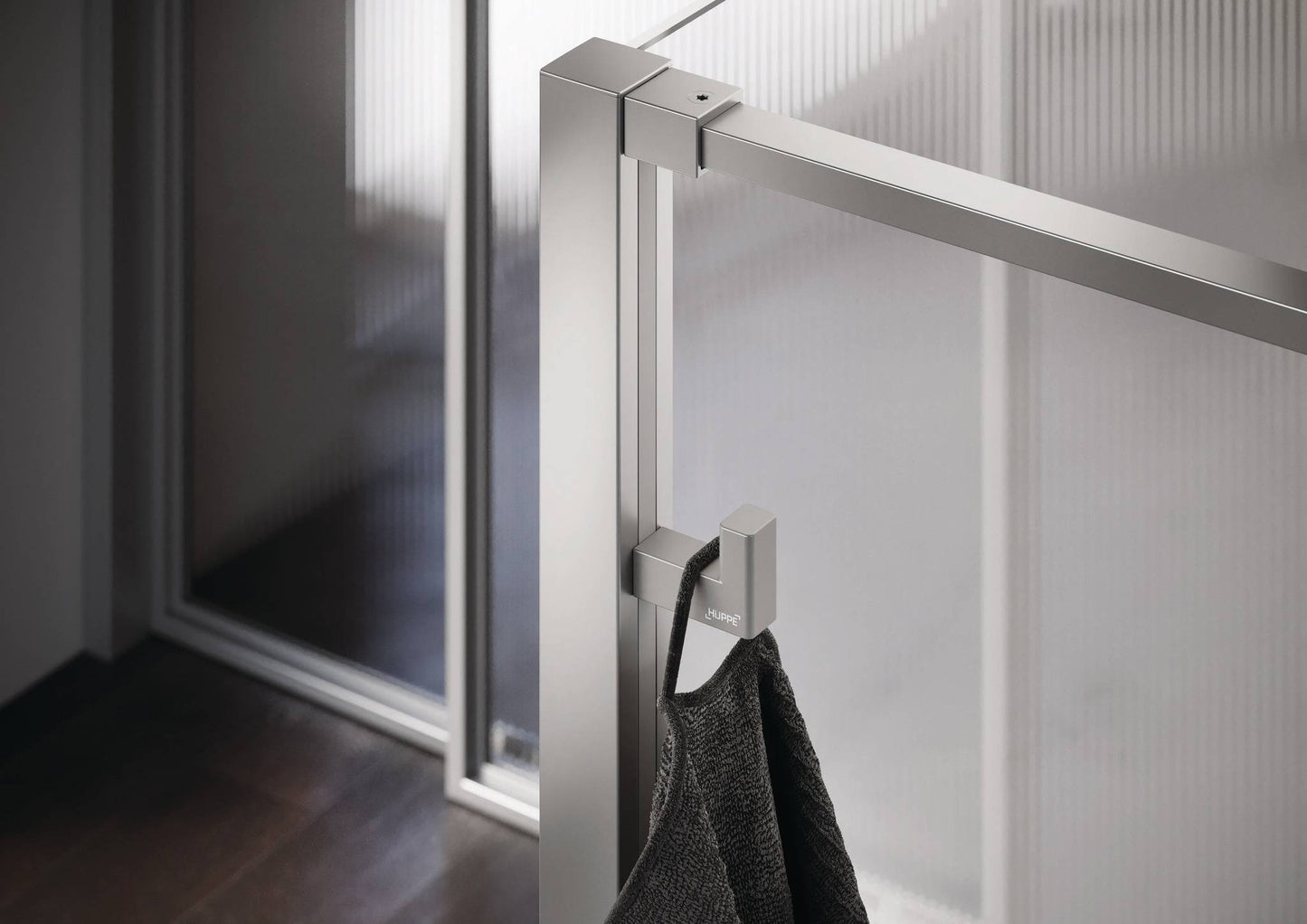 Select+ Hook, silberner Haken der Marke HÜPPE, der an einem senkrechten Profil einer Glasdusche montiert ist, daran hängt ein graues Handtuch, in einem modernen Badezimmer mit dunkelbraunem Fußboden.