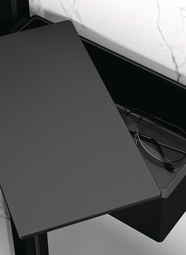 Schwarze matte Select+ Drybox der Marke HÜPPE befestigt an einer Wandleiste einer Glasdusche, mit schwarzem, offenstehendem Deckel, darin liegt eine Brille, vor einer hellen, strukturierten Wand.