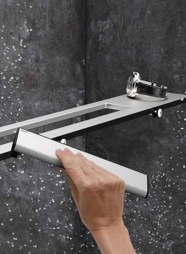 Hand wischt Wassertropfen von einer Glasdusche mit dem HÜPPE Select+ Wiper in silber, während eine Uhr auf einer modernen Ablage vor einer dunklen, strukturierten Wand liegt.