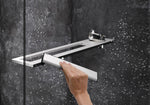 Hand wischt Wassertropfen von einer Glasdusche mit dem HÜPPE Select+ Wiper in silber, während eine Uhr auf einer modernen Ablage vor einer dunklen, strukturierten Wand liegt.