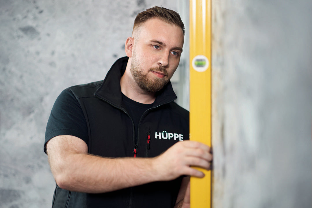 Servicetechniker der Firma HÜPPE misst mit gelber Wasserwage Toleranzen im Duschbereich auf, vor einem grauen strukturierten Hintergrund.