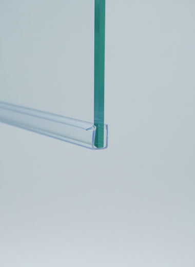 Nahaufnahme einer Ablaufleiste der Marke HÜPPE, transparentes, schmales, längliches, U-förmiges Kunststoffprofil, mit einem Vorsprung an der rechten Profiloberkante, montiert an der Unterseite einer Duschkabine, vor weißem Hintergrund, Seitenansicht links.

