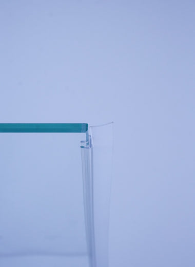 Nahaufnahme einer Anlagendichtung der Marke HÜPPE, transparentes, schmales, längliches, U-förmiges, Kunststoffprofil, mit einem Ablaufschutz an der Profilvorderseite, montiert an Glasdusche, vor hellem Hintergrund, Seitenansicht links.
