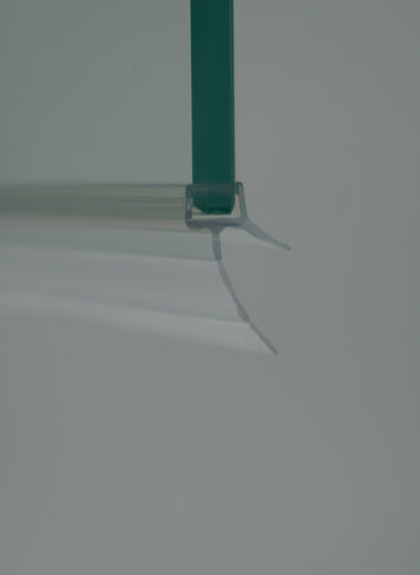 Nahaufnahme einer Ablaufleiste der Marke HÜPPE, transparentes, schmales, längliches, U-förmiges Kunststoffprofil, mit einem Ablaufschutz an der Profilunterseite, montiert an der Unterseite einer Glasdusche, vor weißem Hintergrund, Seitenansicht.
