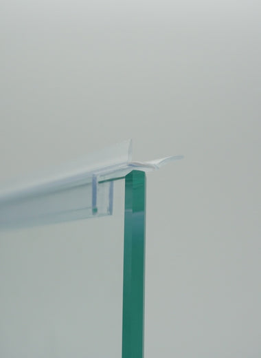 Nahaufnahme einer Ablaufleiste der Marke HÜPPE, transparentes, schmales, längliches, U-förmiges Kunststoffprofil, mit einem Ablaufschutz an der linken und rechten Profilunterseite, montiert an einer Duschkabine, vor weißem Hintergrund Seitenansicht links.
