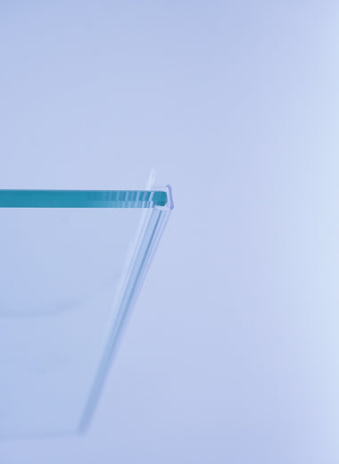 Nahaufnahme einer Anlagenleiste der Marke HÜPPE, transparentes, schmales, längliches, Kunststoffprofil, im Querschnitt P-förmig, mit einem Ablaufschutz an der Profilvorderseite, montiert an Glasdusche, vor weißem Hintergrund, Seitenansicht links.
