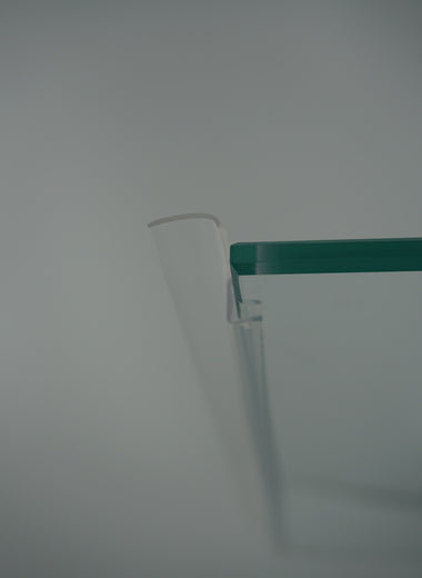 Nahaufnahme einer Anlagenleiste der Marke HÜPPE, transparentes, schmales, längliches, U-förmiges Kunststoffprofil, mit einem Ablaufschutz an der Profilvorderseite, montiert an Duschkabine, vor weißem Hintergrund.

