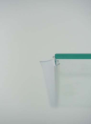 Nahaufnahme einer Anlagendichtung der Marke HÜPPE, transparentes, schmales, längliches, U-förmiges, Kunststoffprofil, mit einem Ablaufschutz an der Profilvorderseite, montiert an Glasdusche, vor weißem Hintergrund, Seitenansicht rechts.
