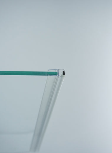 Nahaufnahme einer Magnetleiste der Marke HÜPPE, montiert an Glasdusche, transparentes, schmales, längliches, U-förmiges Kunststoffprofil, mit Magnetprofil an der Vorderseite, vor weißem Hintergrund, Seitenansicht links.
