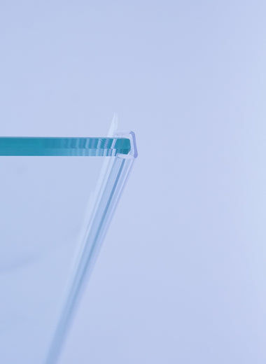 Nahaufnahme einer Anlagenleiste der Marke HÜPPE, transparentes, schmales, längliches, Kunststoffprofil, im Querschnitt P-förmig, mit einem Ablaufschutz an der Profilvorderseite, montiert an Glasdusche, vor weißem Hintergrund, Seitenansicht links.
